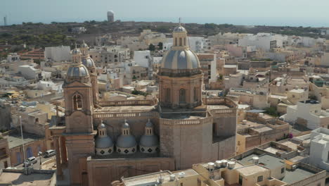 Hermosas-Torres-De-Iglesia-Azules-Y-Marrones-En-Una-Pequeña-Ciudad-Mediterránea-En-La-Isla-De-Malta,-Tobogán-Aéreo-Justo-A-La-Luz-Del-Atardecer