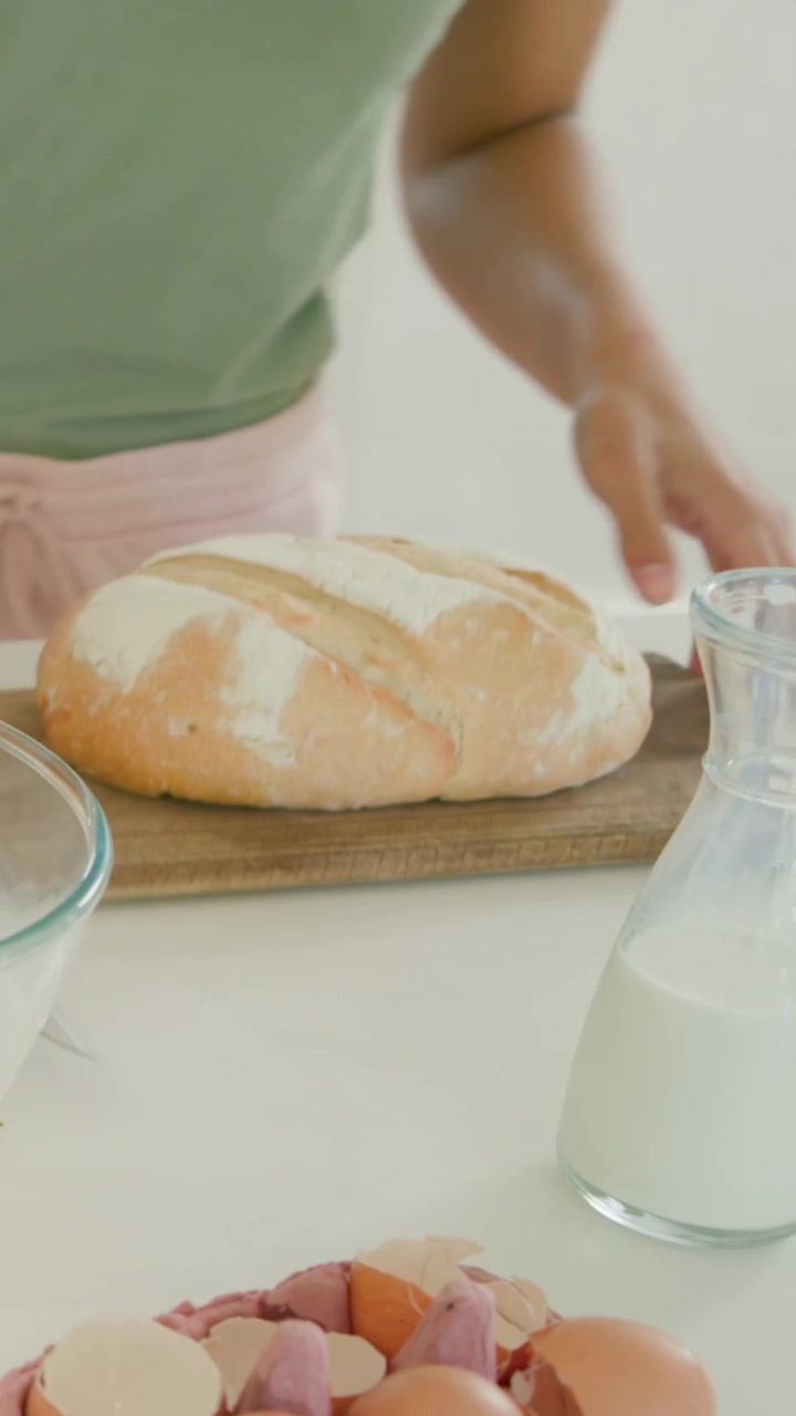 Vertical video: Whisking woman in green top pink shorts, mixing eggs in bowl slicing loaf at home