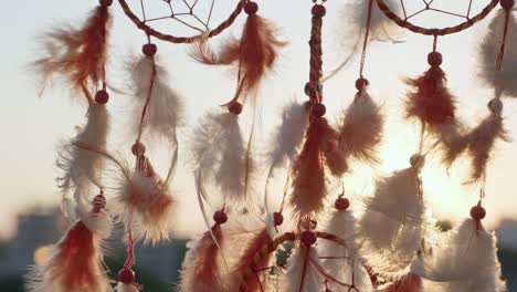 dreamcatcher with orange feathers rotate, swaying with light wind slow motion against background of evening city buildings, blue sky at sunset summer. boho style, indian style. bright disk of sun