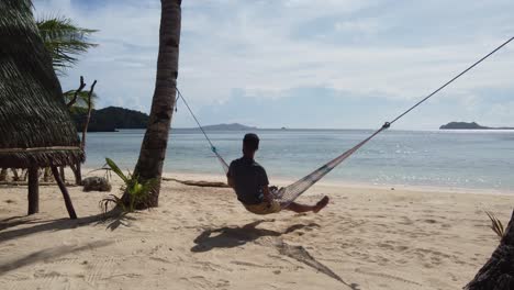 columpio adulto joven en hamaca de árbol en la playa de arena soleada tropical al lado del bungalow