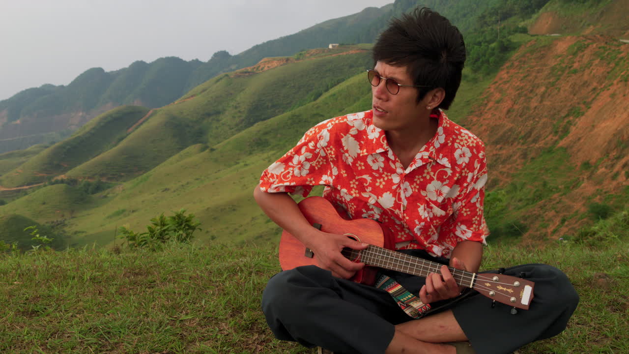 Close up shot of an Asian man sitting on grass while on a trip on top on a mountain and playing ukulele on a windy morning