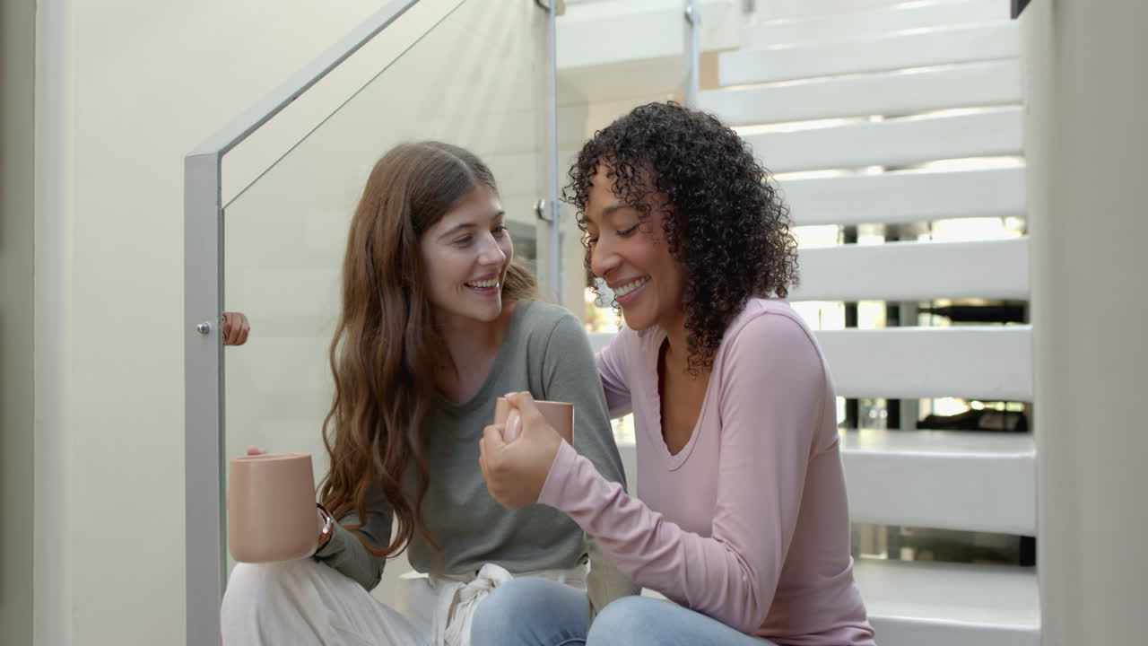Sitting on stairs, lesbian couple enjoying coffee and smiling at home