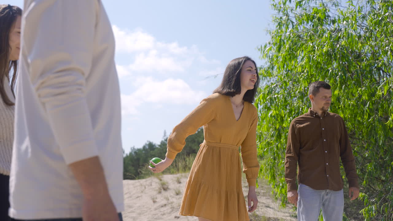 vista lateral de una joven caucásica lanzando una pelota de petanca en la playa en un día soleado mientras sus amigos esperan su turno