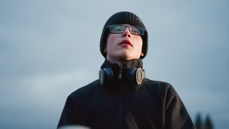 a close-up of a young boy wearing glasses and a black outfit with a headset around his neck, with a blur view of tree in the background