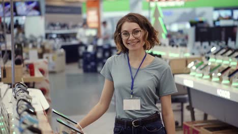 Positive-female-seller-or-shot-assistant-portrait-in-supermarket-store.-Woman-in-blue-shirt-and-empty-badge-looking-at-the
