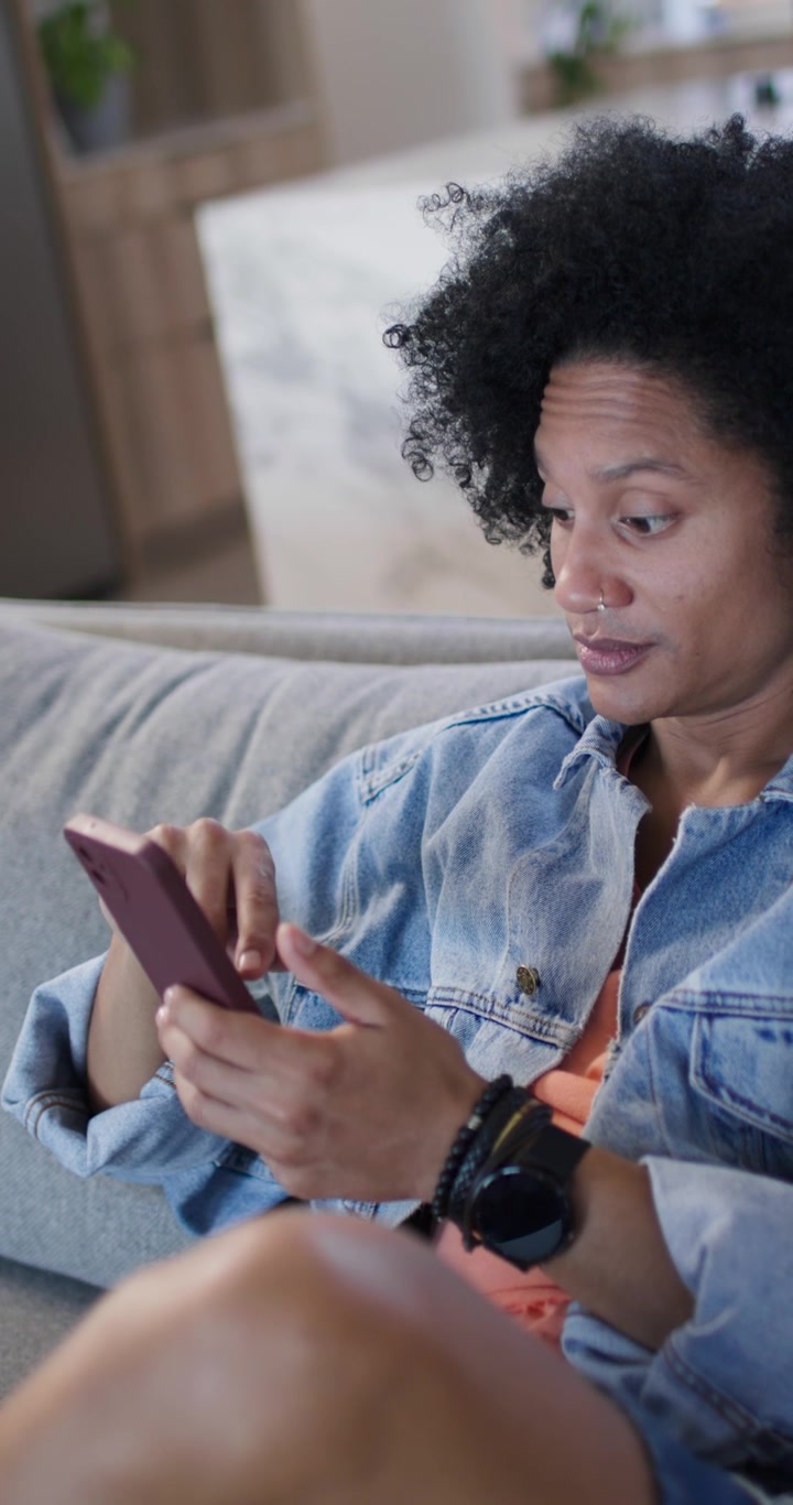 Vertikální video: Afroameričan kontrolující růžový telefon, scrollující na gauči v džínách a čtoucí zprávy.