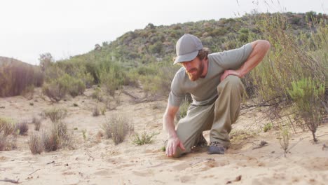 caucasian male survivalist in wilderness squatting down and measuring animal print in sand with hand