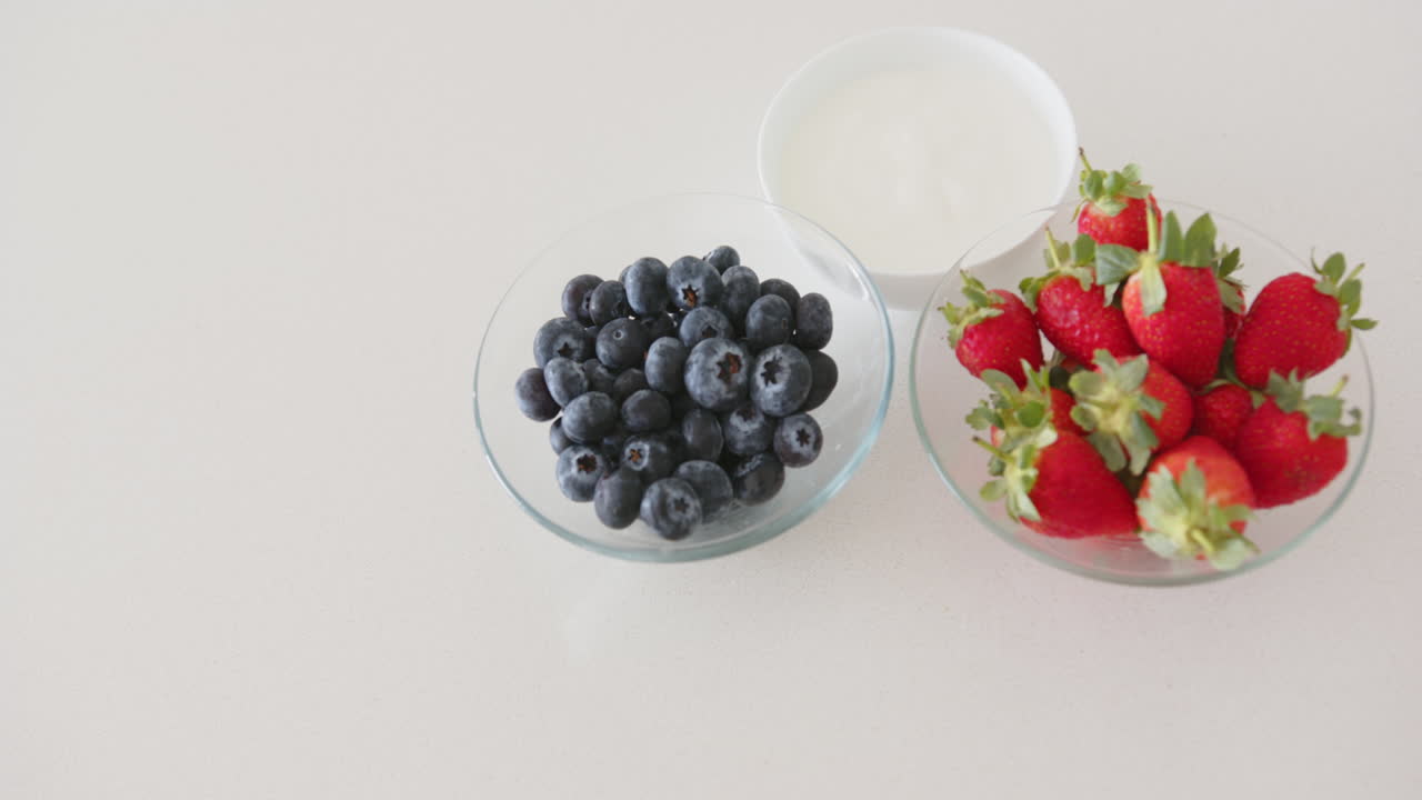 Blueberries and strawberries in glass bowls with yogurt on white surface, at home, copy space