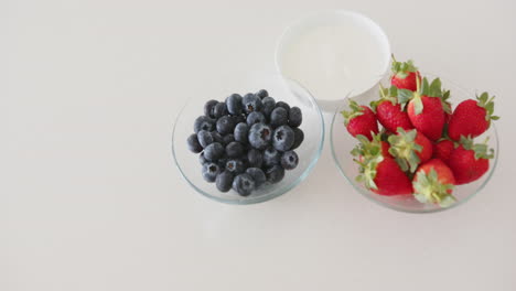 blueberries and strawberries in glass bowls with yogurt on white surface, at home, copy space