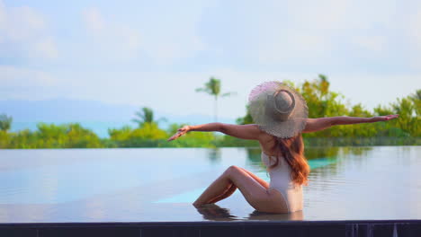 Fit-Asian-woman-up-her-arms-wide-open-while-sitting-at-the-edge-of-the-infinity-pool-with-her-back-to-the-camera-on-tropical-green-background,-daytime-slow-motion
