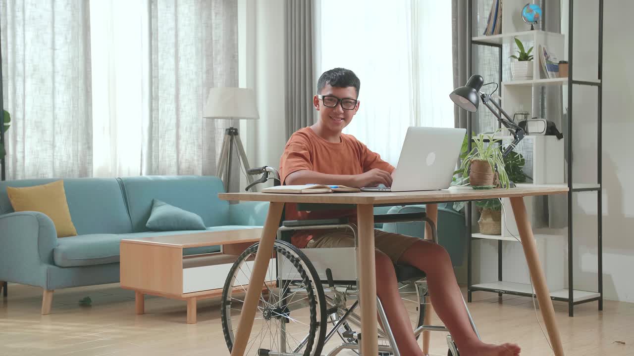 Young Asian Boy Sitting In A Wheelchair Using Laptop Computer While Smiling And Thumb Up To Camera At Home
