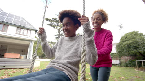 happy african american mother pushing her son on tree swing in garden, slow motion