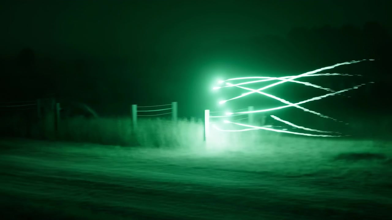 Strisce luminose verde brillante attraversano una scena esterna quasi nera con foschia vicino al centro e deboli sagome sullo sfondo, creando un astratto suggestivo.