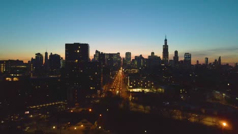 A-captivating-drone-aerial-view-of-a-Chicago-at-dusk,-showcasing-illuminated-skyscrapers-against-the-darkening-sky-The-golden-hues-of-sunset-paint-a-serene-backdrop,-highlighting-architectural-marvels