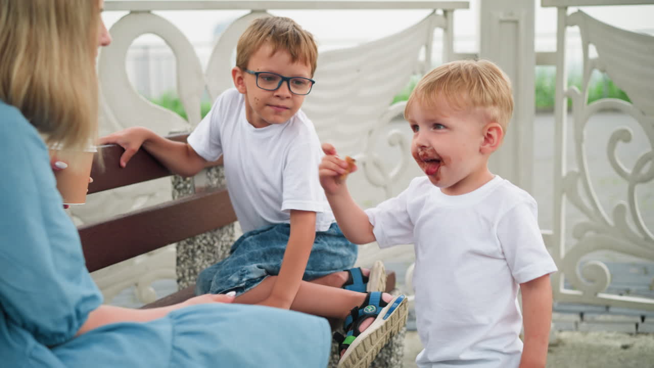 мать и ее двое маленьких детей, младший мальчик, лицо покрытое шоколадом, предлагает свой мороженое своей матери, в то время как старший ребенок смотрит
