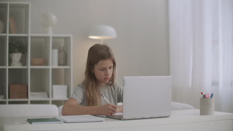 Una-Colegiala-Está-Aprendiendo-En-Línea-En-Casa-Escribiendo-En-Un-Cuaderno-Haciendo-Dictados-Escuchando-A-Un-Maestro-Mediante-Una-Computadora-Portátil-Educación-A-Distancia-Para-Estudiantes