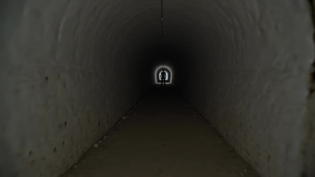Stepping forward dark-clad man toward camera in arched tunnel from bright halo exit with conduit