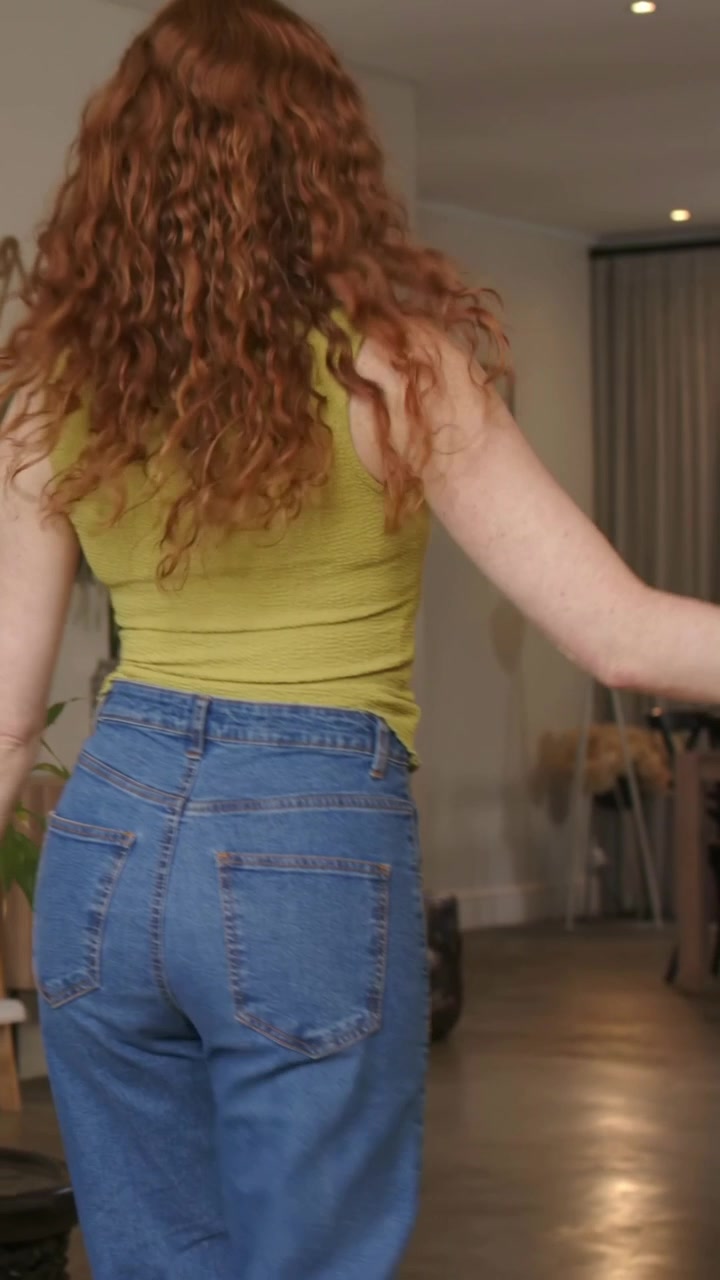 Video verticale: Donna con canotta verde e jeans che entra da sinistra, camminando verso l'ingresso di casa, oltre le piante.