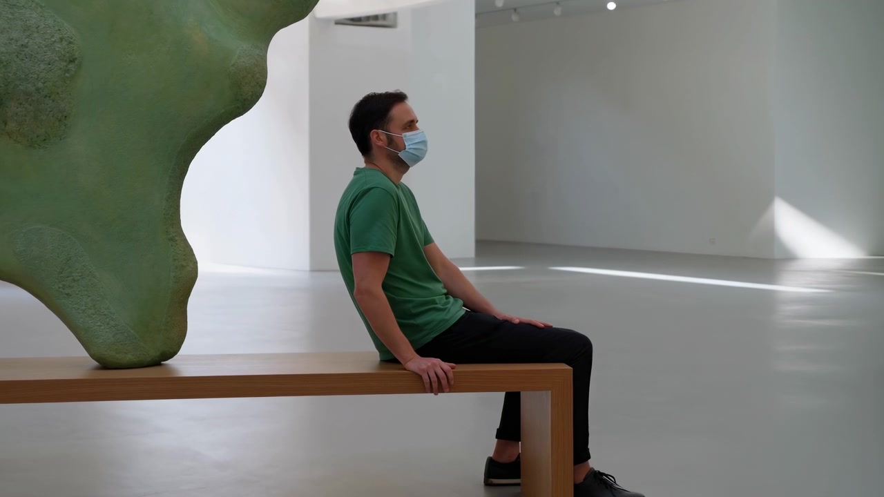 Visitatore mascherato con maglietta verde e pantaloni scuri siede di lato su una panca di legno accanto a una grande scultura astratta verde all'interno di una luminosa galleria bianca, con le mani appoggiate, in uno spazio aperto e tranquillo con luce naturale soffusa.