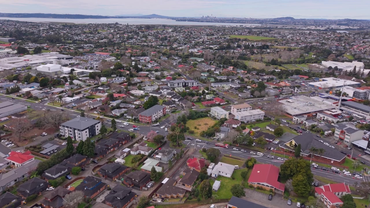 Riprese aeree con drone che sorvolano Henderson, Auckland, mostrando quartieri residenziali, edifici commerciali, strade e il più ampio paesaggio urbano che si estende verso le Waitākere Ranges e la costa.