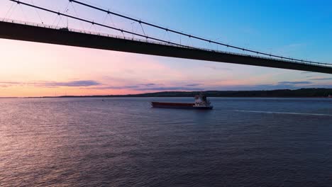 A-tranquil-scene-unfolds-as-a-barge-boat-glides-serenely-under-the-Humber-Bridge-during-sunset,-casting-a-warm,-golden-hue-upon-the-shimmering-river,-captured-in-breathtaking-aerial-footage