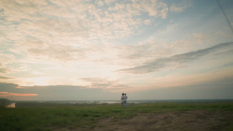 a back shot of a couple standing on a grassy hill beside a lake during sunset. the man, dressed in a white shirt, hat, and jeans, holds the woman by the waist. she wears a black hat and a white dress