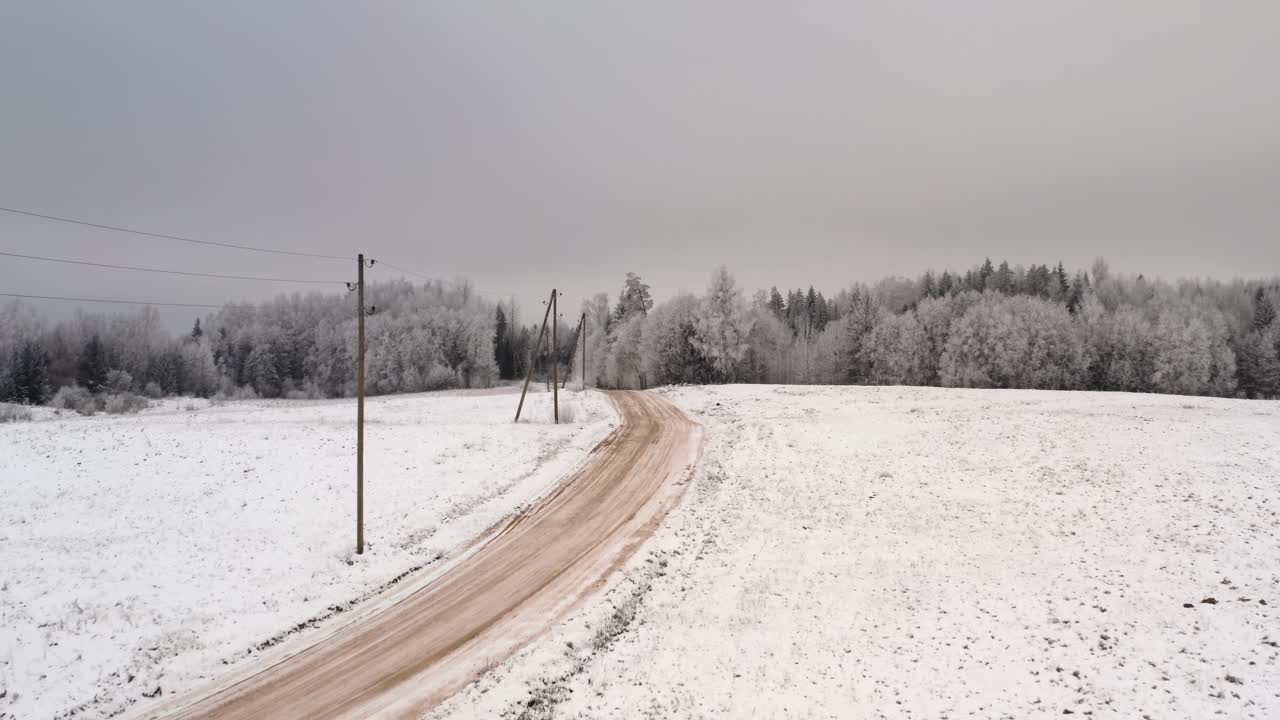 Drone shot of gravel road in winter season. Aerial winter day scenery with snow and frost covered forest. Power lines along the road with wooden poles.