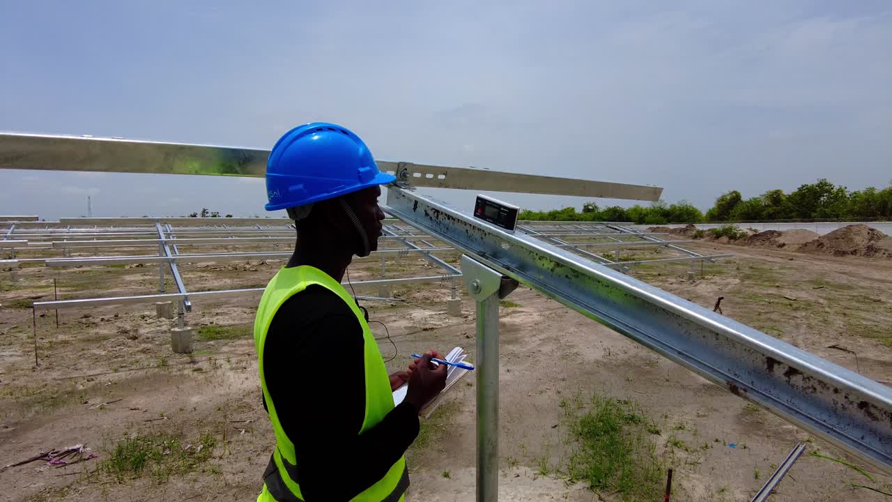 Black African engineer wearing blue helmet taking measurements of solar panel array beam structure tilt angles