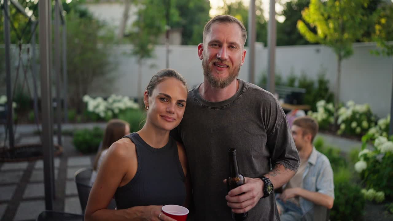 Portrait of a happy blond guy with a beard in a gray T-shirt who hugs his girlfriend and poses while relaxing with company in the courtyard of a country house