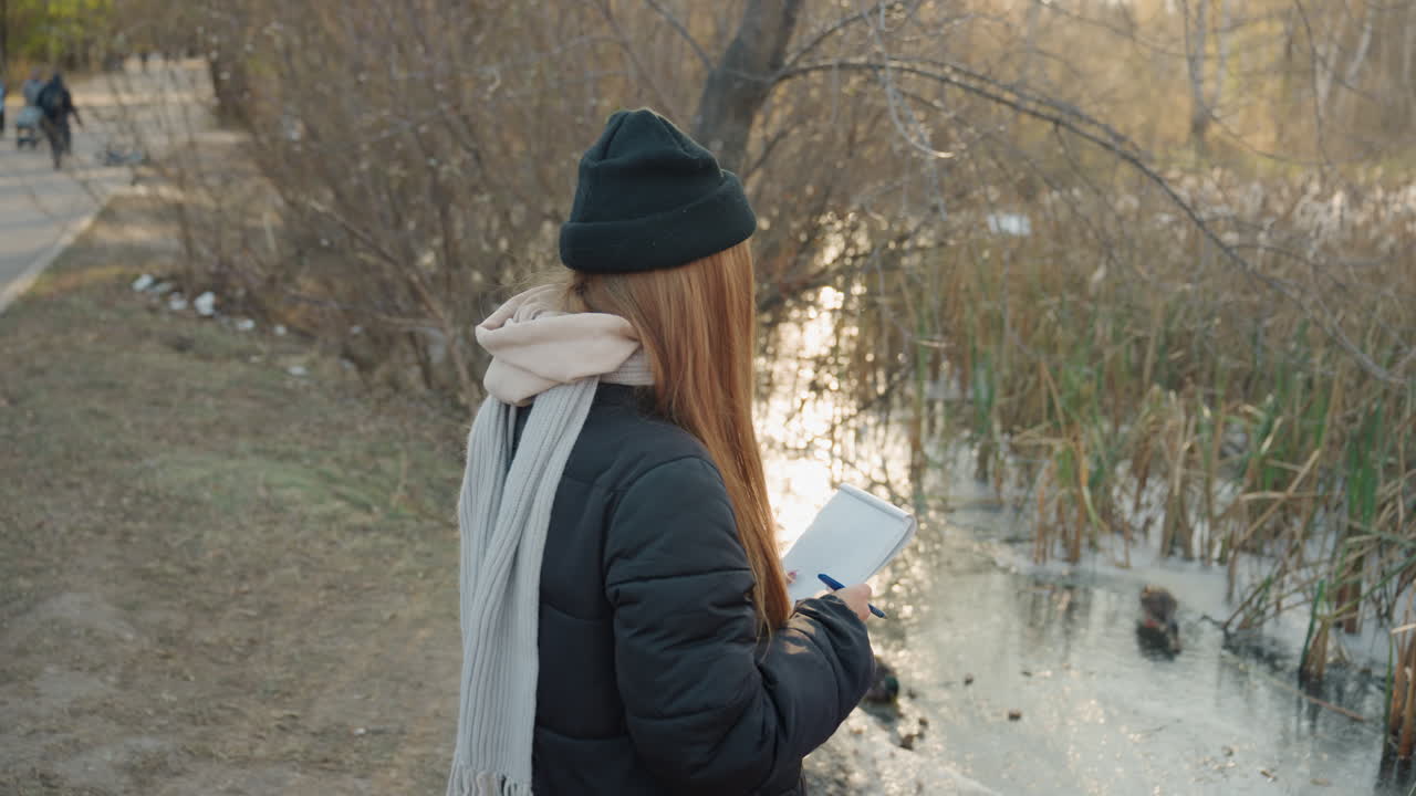 Young woman by pond at dusk holding phone and notebook, braided hair with black beanie and scarf, walking along water edge, quiet autumn light, reflective mood, student checking messages, writer