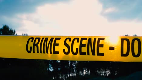 close up footage of a yellow police investigation forensic crime scene tape that is pulled infront of sunset blue sky with white clouds and dark forest treeline visible also. tape is backlit by sun