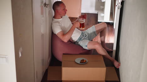 remote worker seated casually on soft beanbag chair enjoying break, holding meat in one hand and glass mug of fruit juice in other, placing mug back on cardboard box with plate beside him in hallway