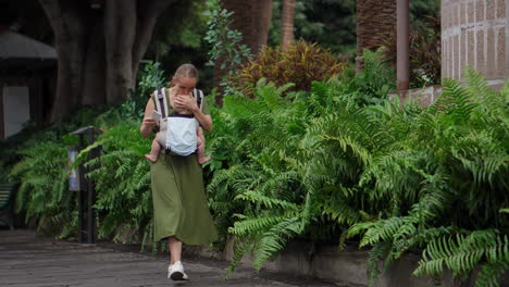 while using a kangaroo backpack to carry her baby, a young woman snaps pictures on a mobile phone during her travels. she walks and checks the phone screen at times