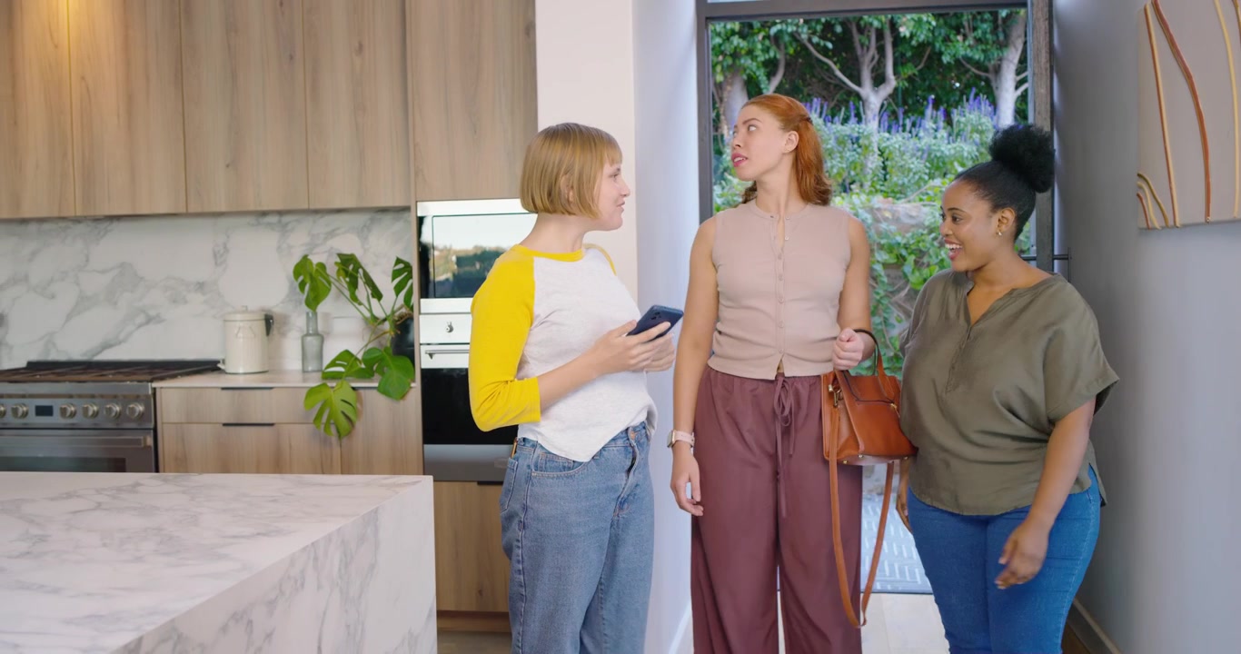 Diverse female friends entering front door, hugging, inspecting kitchen island, checking smartphone