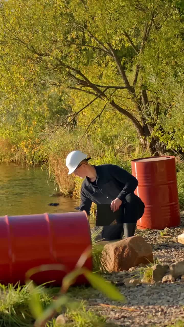 Man collects water sample from lakeshore while kneeling on ground between oil drums, vertical