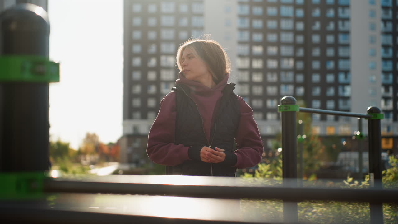 Outdoor trainer in hoodie and vest warming up by exercise rod bar under sunshine, calmly rolling hands then turning head sideways with focused expression while people stroll in urban park backdrop