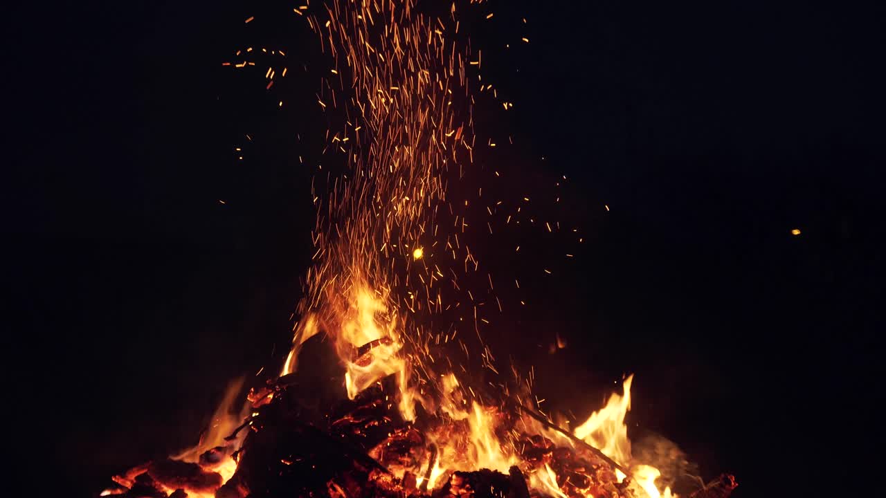 Burning fire with logs and coals at night. Bright sparks and heavy smoke from the fire. A man turns firewood with a pitchfork.. Burning branches in the fire, tongues of flame