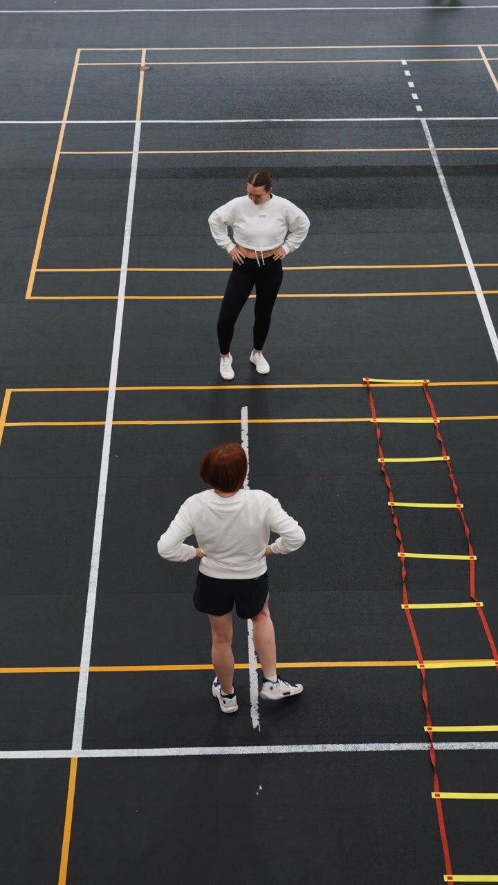 Intenso esercizio di gioco di gambe per il badminton in campo, allenatore e giocatore si esercitano con agilità con la scaletta e passi laterali, riscaldamento mirato, ritmo misurato e ripetizione, linee minimaliste della palestra interna e pavimento scuro, dall'alto