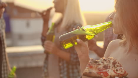 a women eat piece of hot pizza and drink beer from a green glass bottle on the party with his fried in hot summer day.