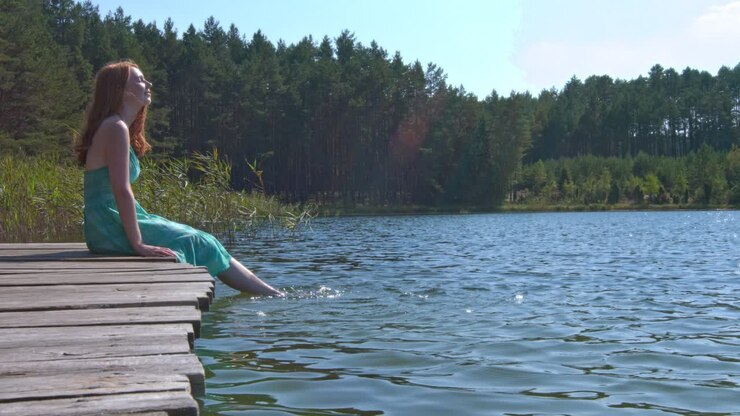 Una donna che si rilassa, seduta sul bordo di un pontile, sguazza i piedi nell'acqua di un lago fluviale.