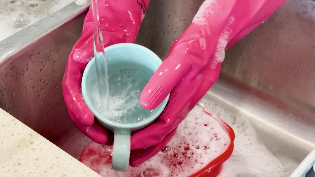 Hands in pink gloves thoroughly wash a blue ceramic mug under running water in a kitchen sink with soapy dishes and bright, even lighting