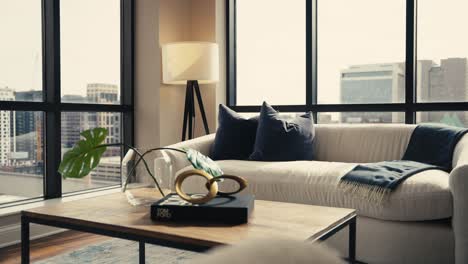 a living room setup next to large windows with a white comfy couch, blue pillows, a coffee table, and the city skyline in the background