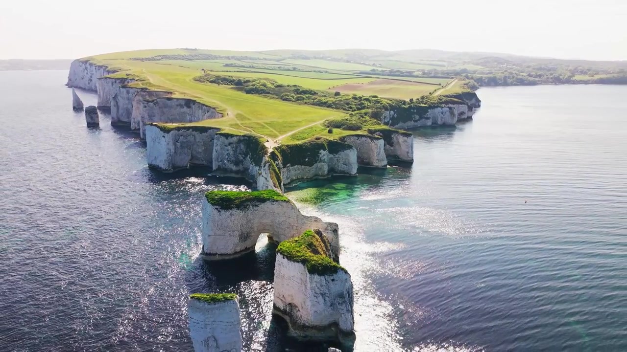 Široký letecký záběr z dronu na Old Harry Rocks na pobřeží Dorsetu v Anglii, ukazující bílé křídové útesy, skalní útvary v moři, přírodní mořský oblouk, tyrkysovou vodu a zelené stezky na útesech.