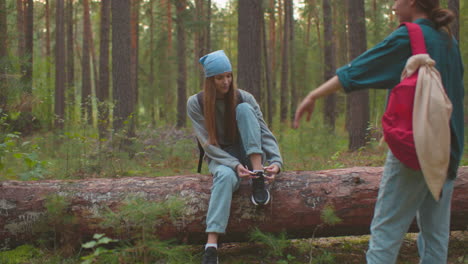 friends take a break in a forest, one sitting on a fallen tree tying her shoe, while her companion stands nearby with a red backpack, engaging in conversation, tall trees and greenery surround them