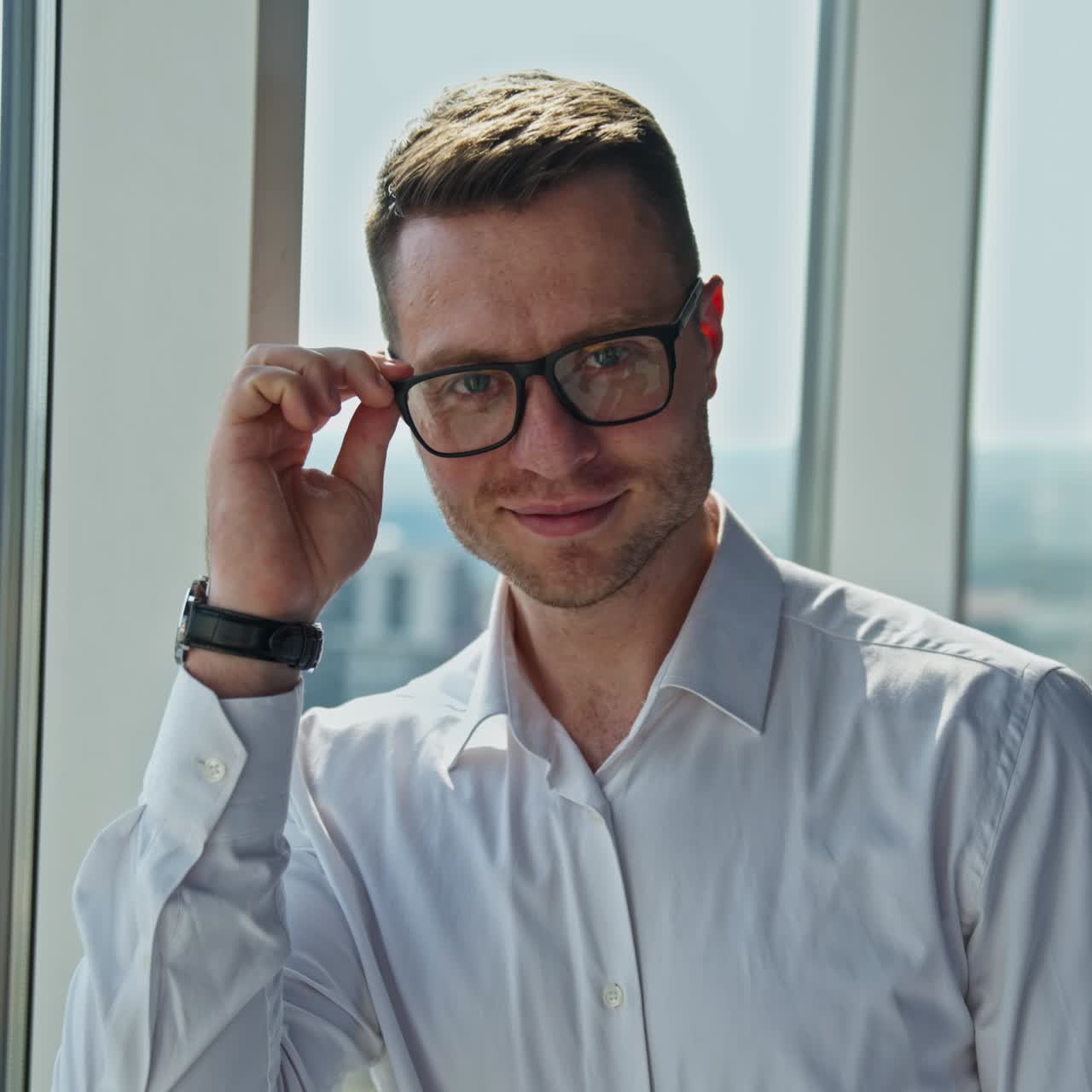 Thoughtful mid-aged man stands leaning on panoramic window. Man puts on glasses looking straight into camera smiling