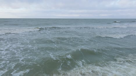 Vista-Aérea-De-Las-Grandes-Olas-Tormentosas-Rompiendo-Contra-La-Playa-De-Arena-Blanca,-Día-Nublado,-Dunas-Costeras-Dañadas-Por-Las-Olas,-Erosión-Costera,-Cambios-Climáticos,-Amplio-Tiro-Bajo-De-Drones-Avanzando