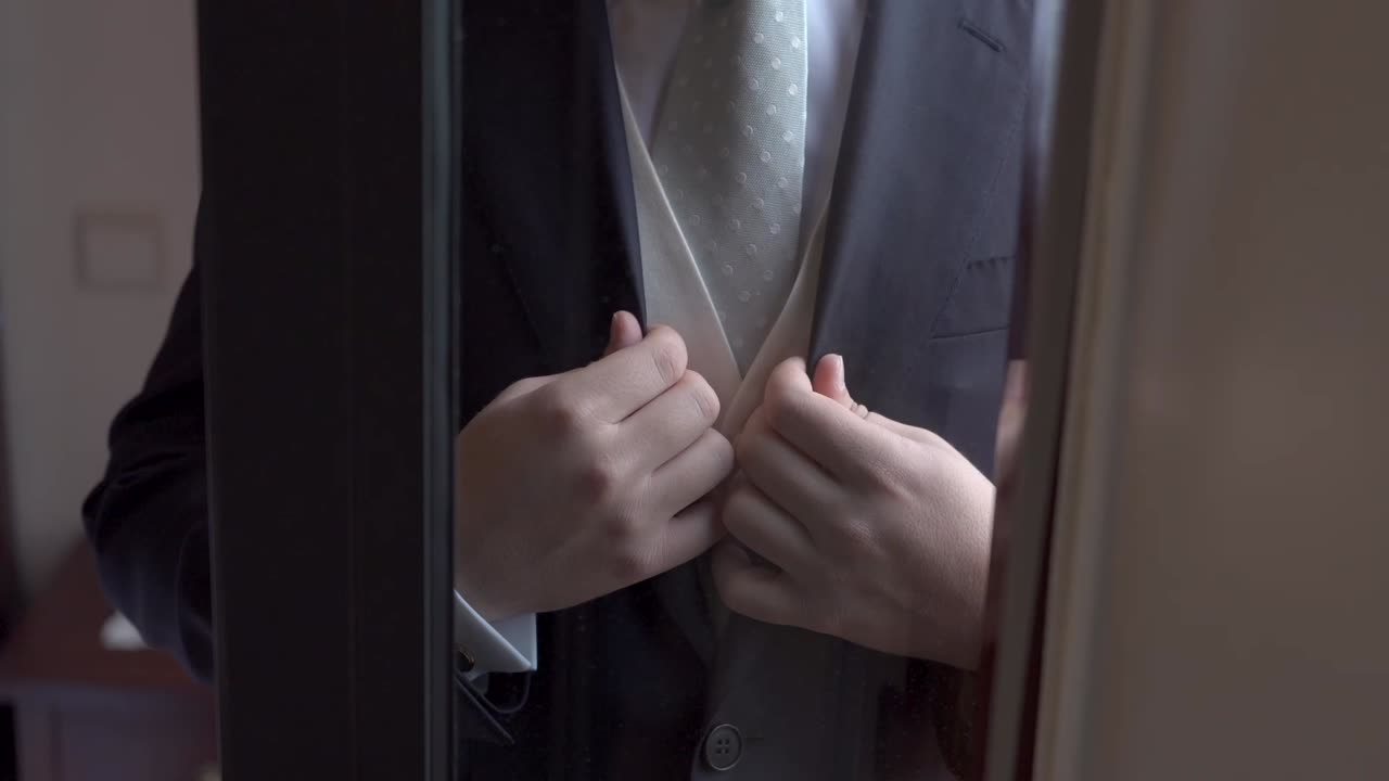 Groom fixing his dark suit and white tie through a window before wedding ceremony