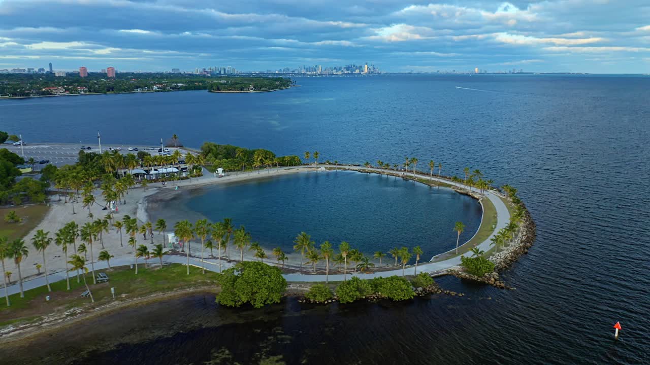 The iconic atoll pool curves along the Biscayne Bay shoreline as palms, mangroves, and distant Miami skyline views frame the coastal landscape of Matheson Hammock Park