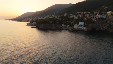 aerial view of italian coastline at sunset behind mountains, golden sky and calm sea surround hillside villas, drone orbits clockwise