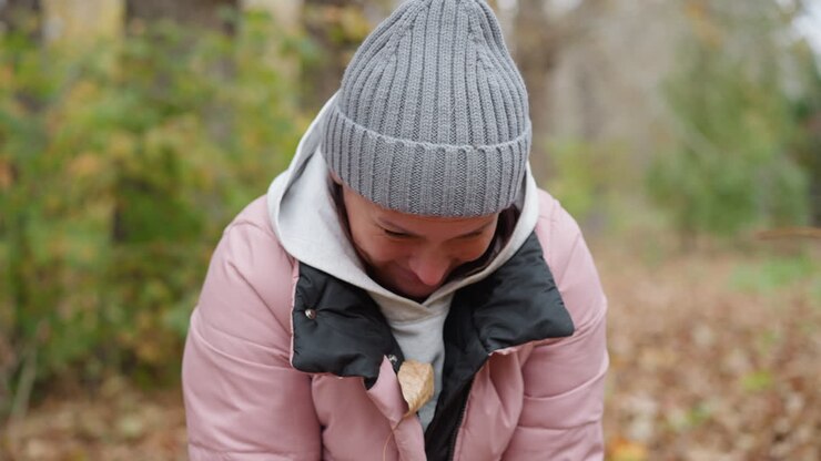 Joyful moment as woman in pink jacket and grey beanie laughs while autumn leaves fall around her, creating a playful and vibrant autumn scene